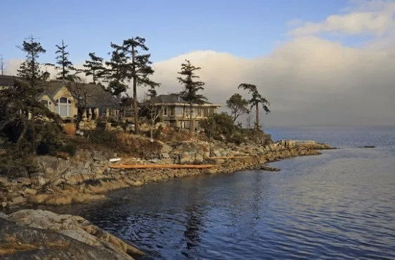 Luxury homes perch atop rocky cliffs on Vancouver Island, overlooking calm waters under a sky with soft clouds, illustrating a serene weather condition.
